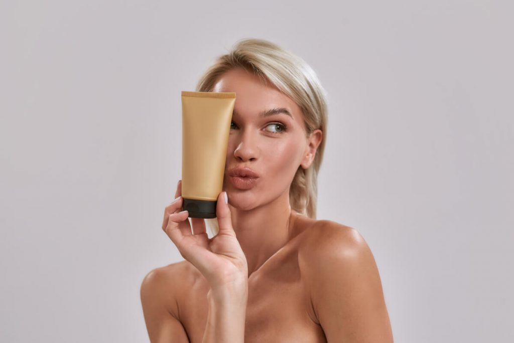 Portrait of cheerful young woman with perfect glowing skin looking aside, holding sunscreen skincare product